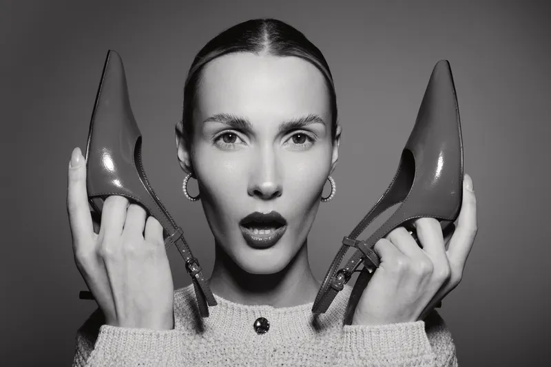 A young white woman with pale skin holds two high heel shoes near her face in a surprised expression, close-up black and white portrait.