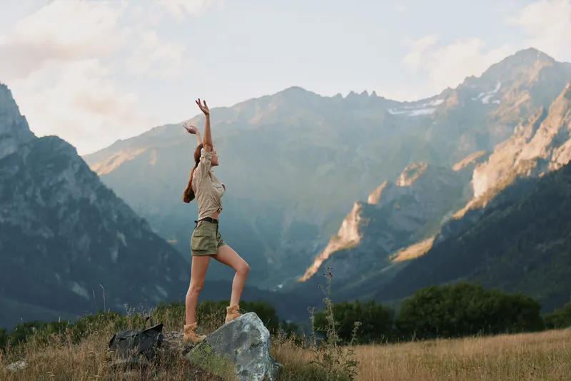 A woman in casual clothes stands on a rock throwing a ball in a mountain landscape. The scene shows active lifestyle and outdoor recreation in nature.