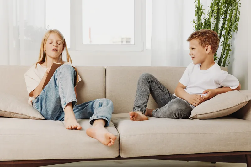 Two children relax on beige sofa in bright living room, girl reading book and boy smiling while sitting barefoot. Concept of childhood, leisure, home comfort, and family time.
