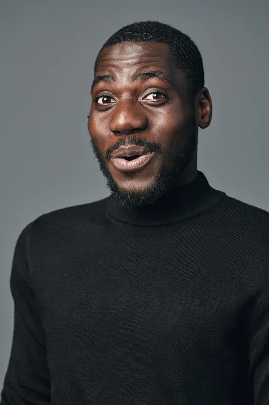 African man with dark skin wearing black turtleneck sweater in creative conceptual visual style looking surprised with open mouth and expressive face on gray background studio portrait.