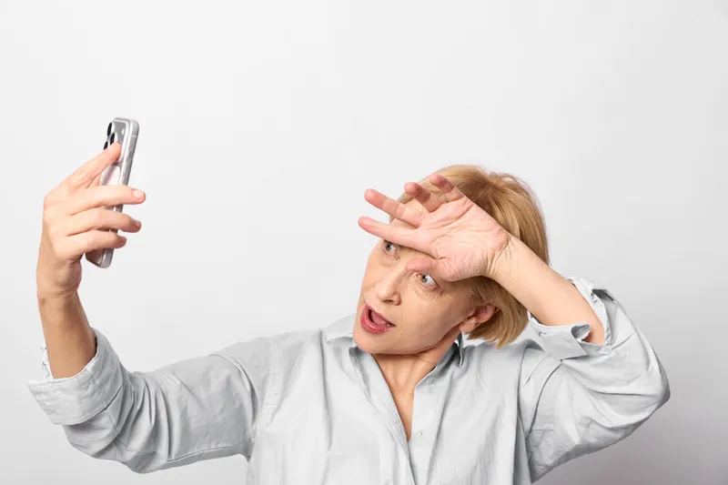 Woman holding smartphone for selfie, covering her forehead and squinting with stress, tired face gesture on a white studio background, gray shirt, soft light, health concept