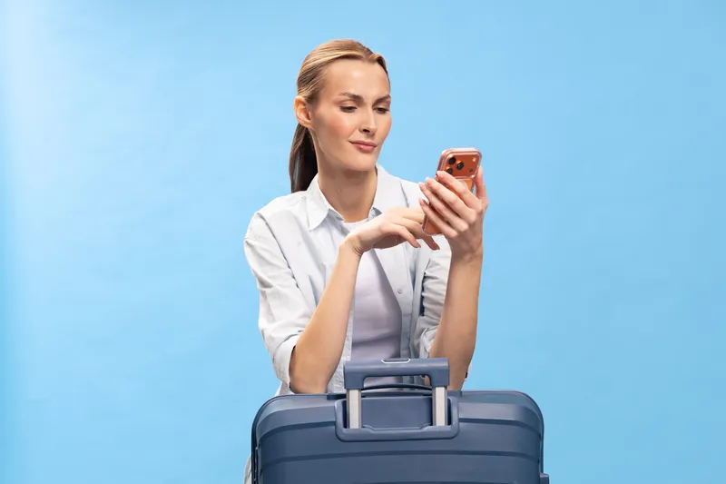 Young woman in white shirt stands behind blue suitcase looking at smartphone with interest on blue background. Concept of travel planning, vacation, and communication.