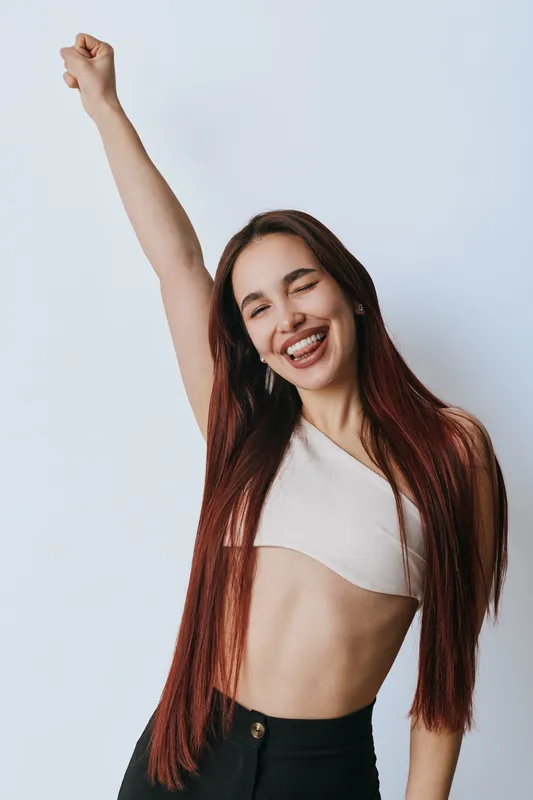 Young woman with long hair smiling and raising fist in celebration, wearing white crop top and black pants against plain background for happiness and confidence.