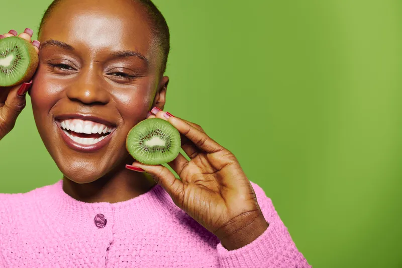 Smiling dark skinned African woman holds two kiwi fruit slices near her face, wearing pink sweater. Green background creates fresh, healthy concept of fruit, skincare and wellness lifestyle.
