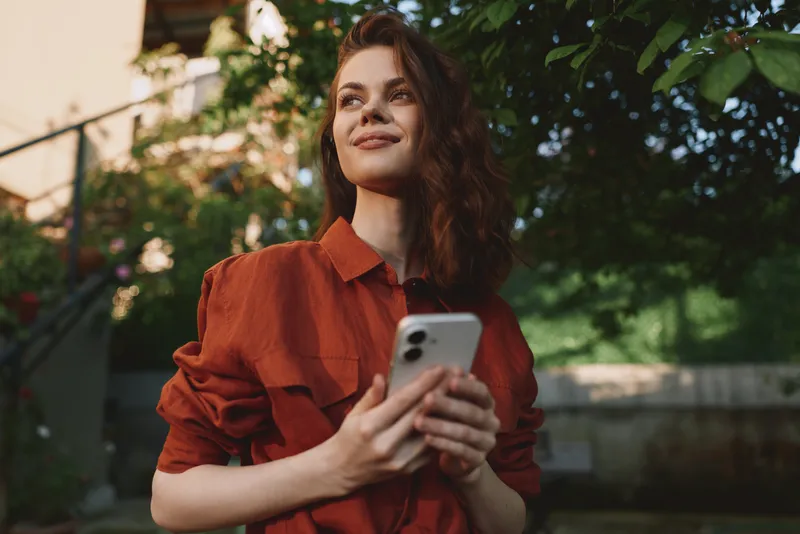 A woman in a red blouse stands outdoors, holding a smartphone with a calm, casual pose. Sunlight and greenery frame a relaxed lifestyle moment