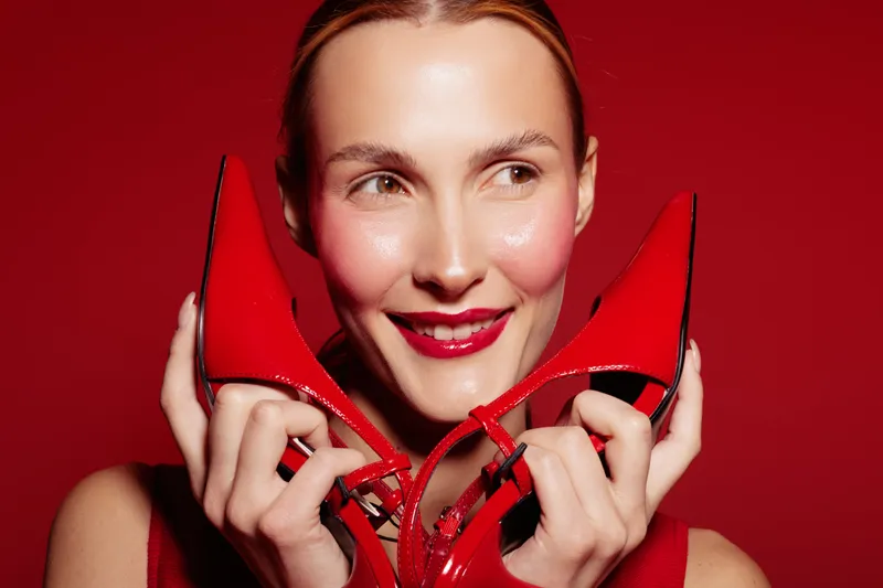 Smiling woman with red lipstick holds red high heel shoes near face on red background. Concept of fashion, beauty, style, and elegance with vibrant color contrast.