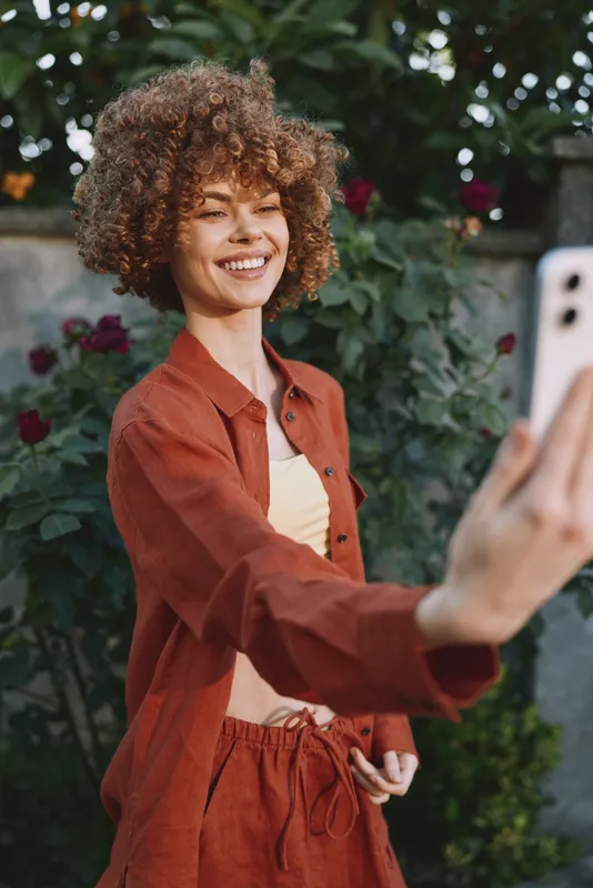Woman lifestyle image of a smiling woman taking a selfie in a garden, wearing casual red clothing, radiating joyful mood and relaxed atmosphere in outdoor light