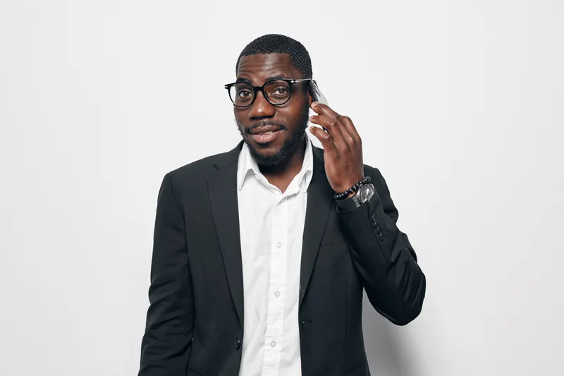 African American man with dark skin wearing glasses and black suit jacket talking on smartphone in studio. Professional male with beard and white shirt using mobile phone for communication.