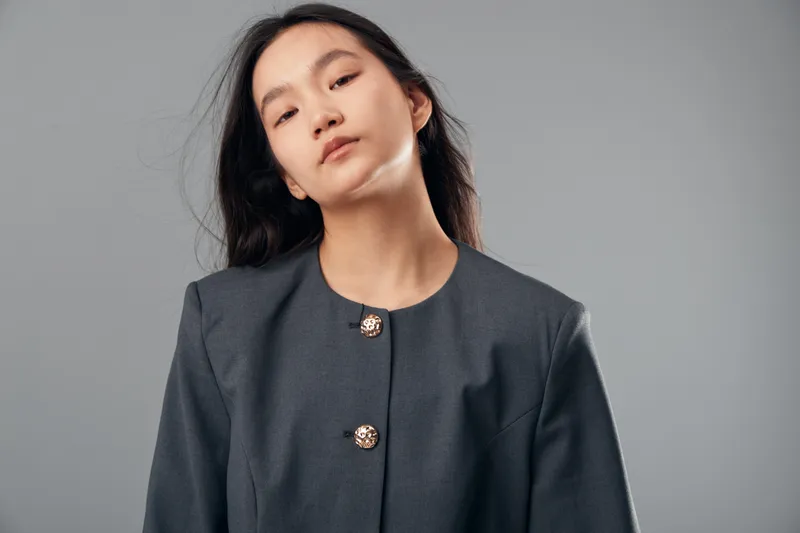 Young woman posing in dark gray jacket with brass buttons against gray background for fashion portrait and confident look in studio lighting and straight hairstyle.