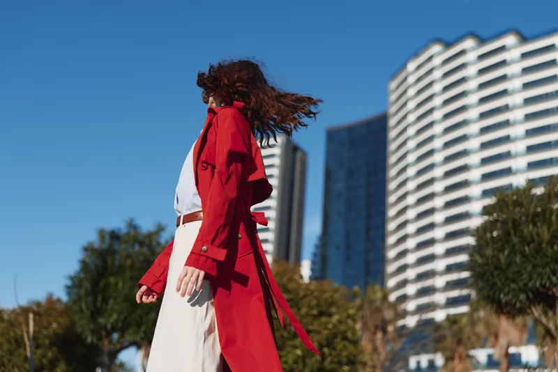 A woman in a red trench coat walks outdoors near modern buildings on a sunny day. Her hair flows in the wind, capturing movement and urban lifestyle.