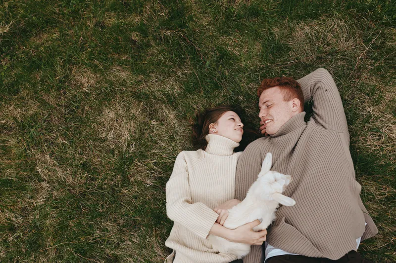 Couple lying on grass sharing a tender moment with a playful goat kid, cozy sweaters, relaxed lifestyle scene, outdoor romance, gentle smiles, natural light, candid family vibe.