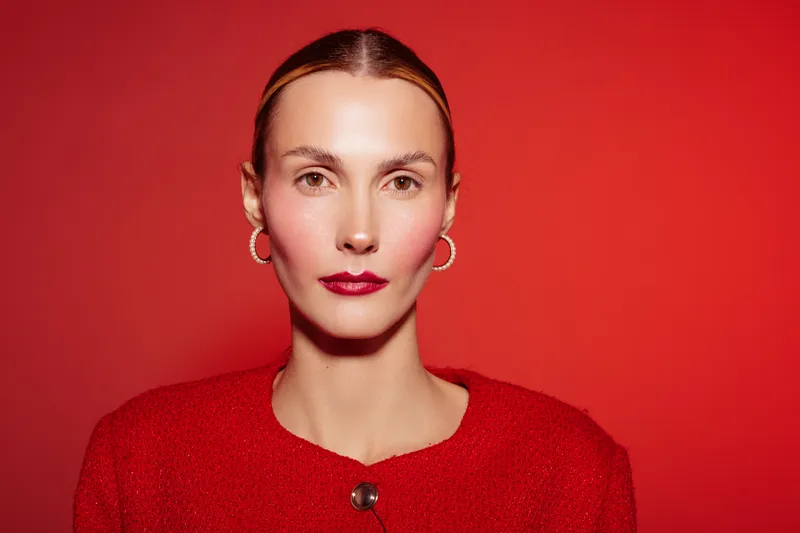 Portrait of confident woman with slicked hair and red lipstick wearing red sweater and hoop earrings against red background. Concept of fashion, beauty, and elegance in studio lighting.
