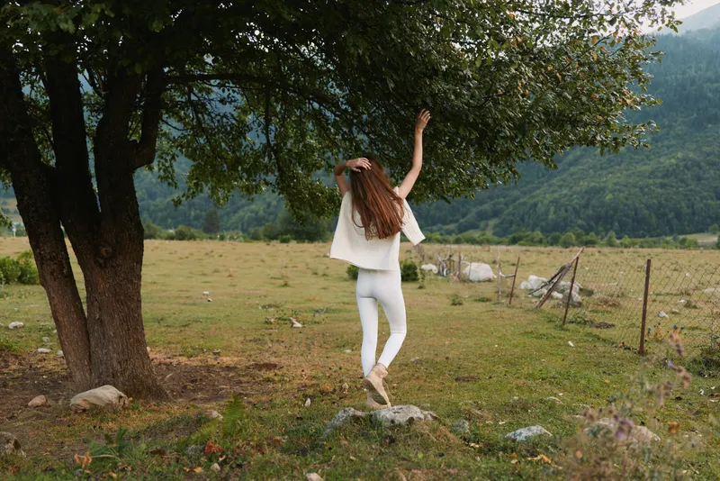 Young woman in white outfit leaps across a grassy meadow beside trees, capturing a carefree moment in nature with fluttering pants and sunlit hills that convey energy and joy.