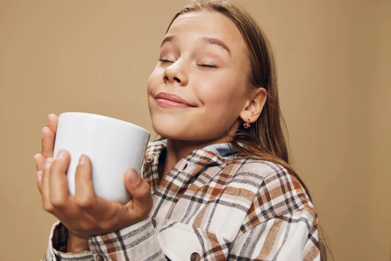 Young woman holding white mug with closed eyes and slight smile wearing plaid shirt on beige background enjoying warm drink and cozy moment indoors.