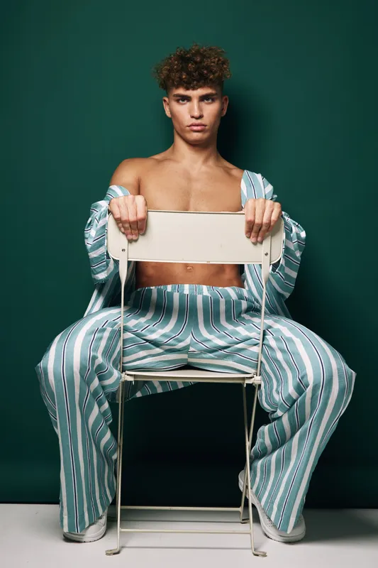 Young man in striped pants and open shirt sits on metal chair backward against green background. Studio portrait of confident male model with curly hair, relaxed pose and barefoot style.