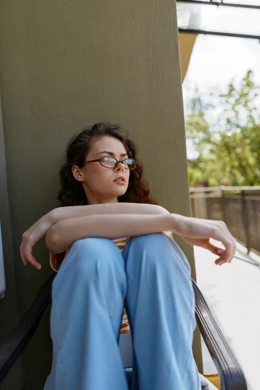 A pensive young woman sits on stairs in daylight, wearing blue scrubs and glasses, arms wrapped around knees. A quiet moment of reflection, isolation, and mental wellness in a calm, intimate setting.