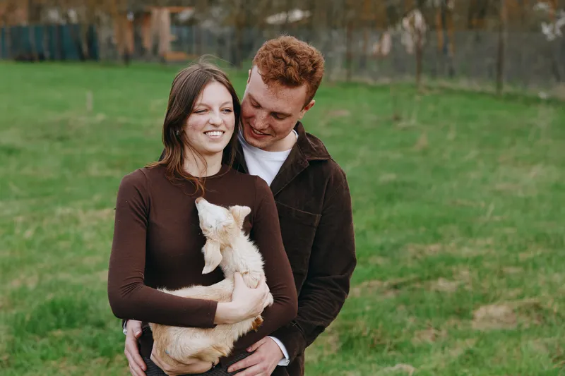 A young couple embraces outdoors while the woman holds a small goat in her arms, sharing a tender moment in a casual lifestyle setting.