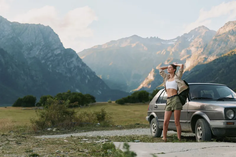 A young woman with a backpack stands beside a car in a mountainous valley, ready for an outdoor hike.