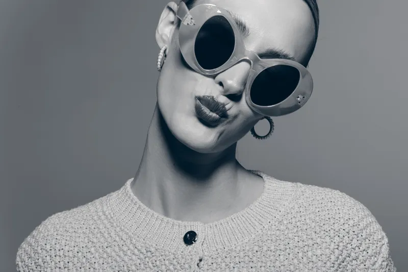 Stylish young caucasian woman with pale skin wearing large round sunglasses and knitted sweater poses with playful lips on grey background.