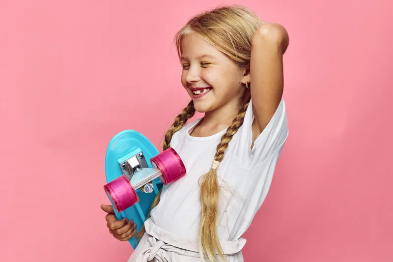 Happy young girl with braided hair holding a blue skateboard with pink wheels smiles with eyes closed against a pink background. Concept of childhood, leisure, and joy.