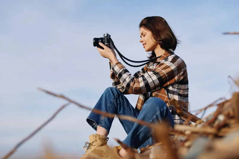 Woman photographer outdoors with a camera, sitting on a grassy hillside, capturing scenery in natural light, casual plaid shirt and jeans, reflecting creativity and travel mood