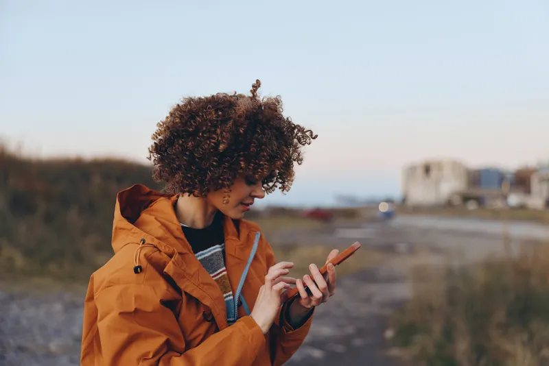 Woman lifestyle with curly hair, wearing orange jacket, outdoors on a street, checking a smartphone, warm sunset light, city buildings blurred, casual travel mood