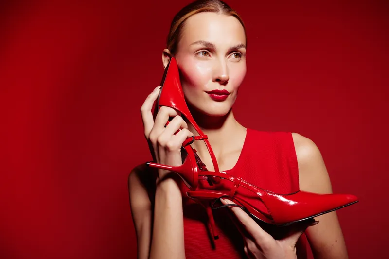 Young woman in red dress holds red high heel shoe near face against red background. Concept of fashion, style, elegance, and beauty with vibrant color contrast and modern look.
