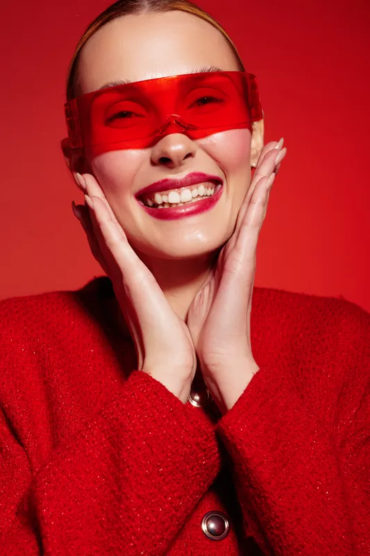 Smiling woman in red coat and red translucent glasses poses with hands near face on red background. Concept of fashion, joy, style, and vibrant color contrast in studio lighting.