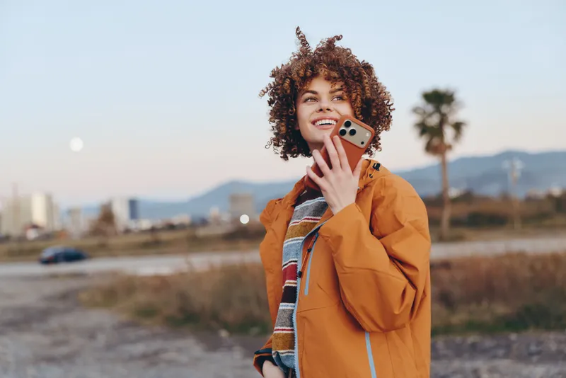 Woman lifestyle outdoors, smiling curly portrait, holding phone to ear for communication call, walking on park path, warm autumn sunlight, mountain landscape, palm trees