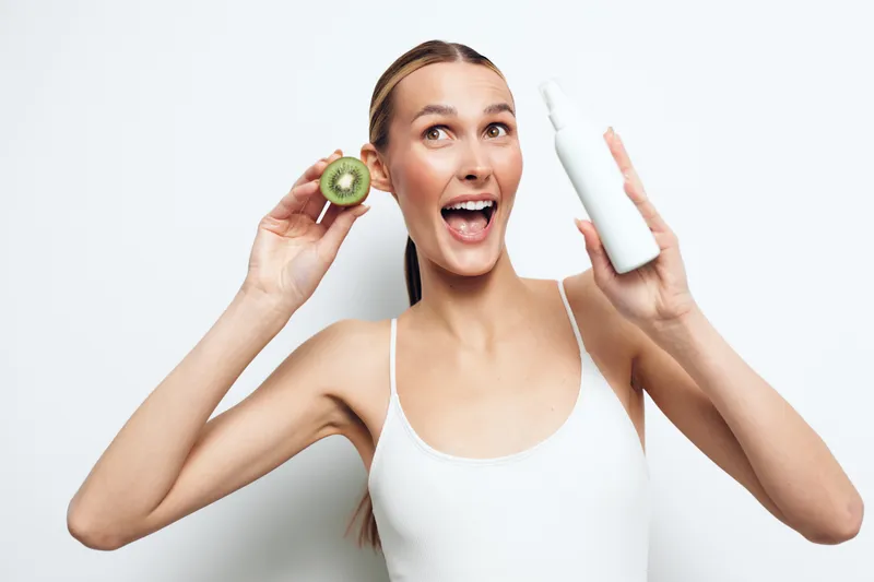 A cheerful young woman in a white tank top holds half a kiwi fruit and a white skincare bottle, smiling with excitement against a plain white background. Concept for beauty and freshness.