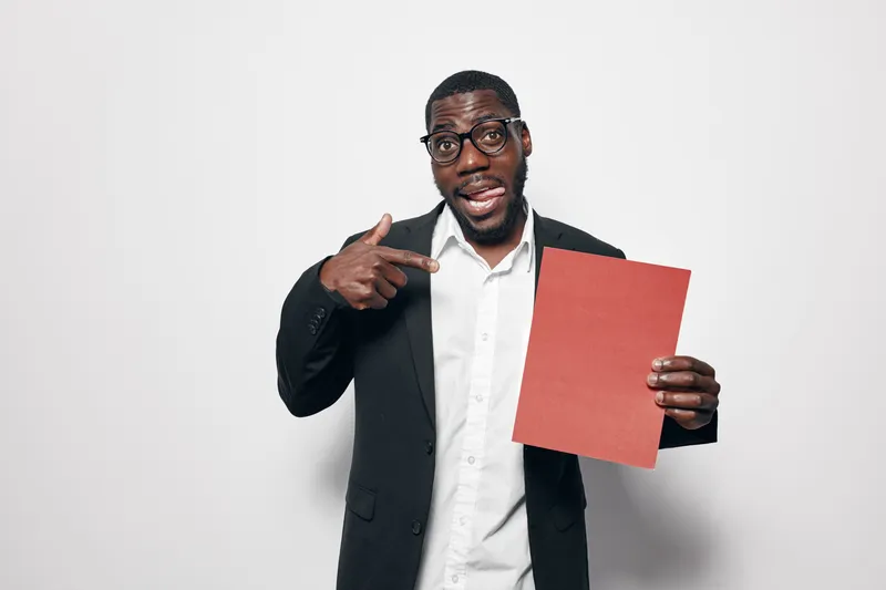 African American man with dark skin wearing glasses and black jacket pointing at red paper sheet on white background. Confident male holding blank document for presentation or advertisement.