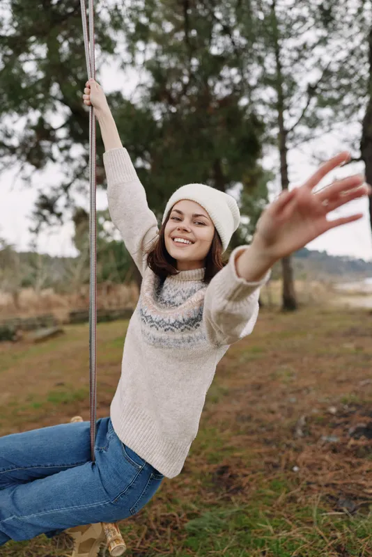 Girl on swing in an outdoor scene, a joyful moment captured in a park setting with trees and open space, wearing a sweater and beanie for a cozy, candid mood.