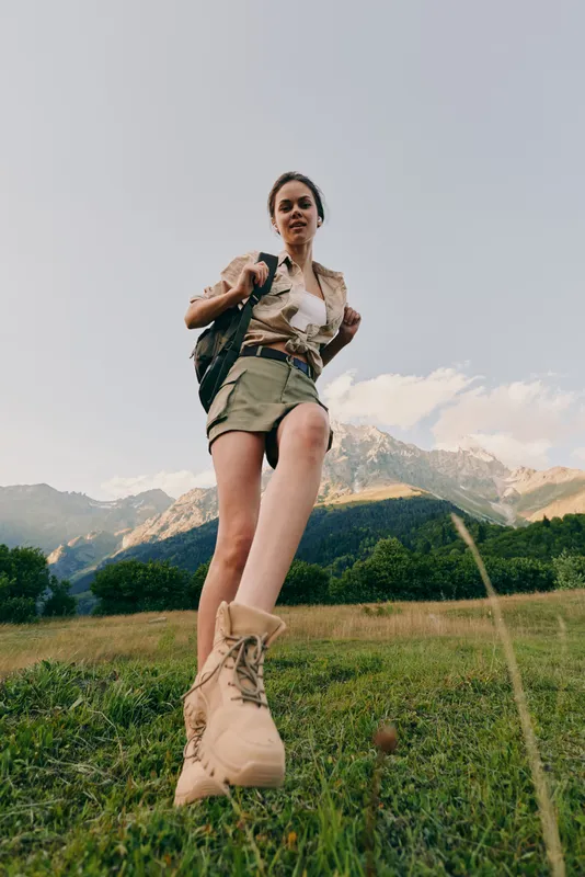 A young woman hikes across a grassy meadow beneath towering mountains, backpack slung over her shoulder, boots crunching the moss as she strides forward with confident, adventurous energy.