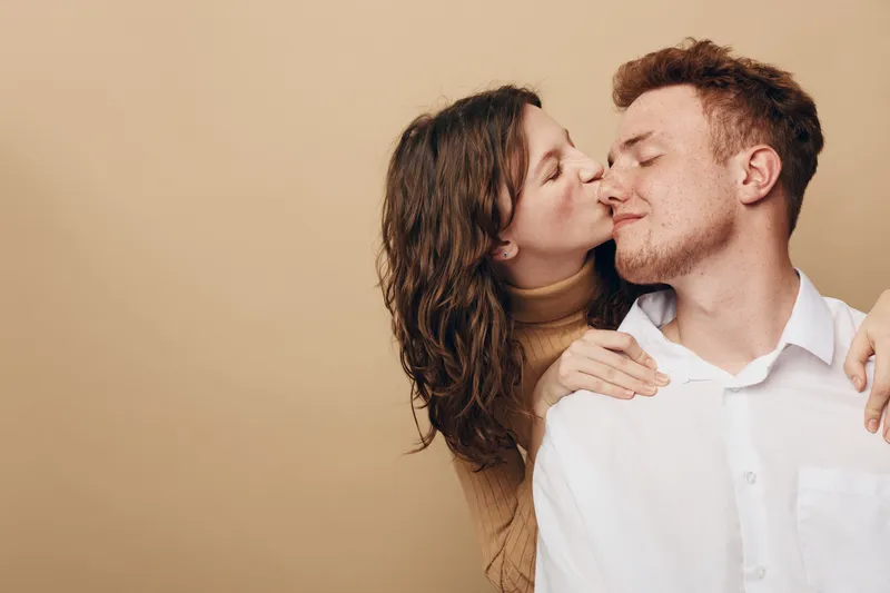 Young couple in casual clothes share intimate moment, woman kissing man on cheek with closed eyes. Warm beige background creates cozy romantic atmosphere and genuine affection concept.