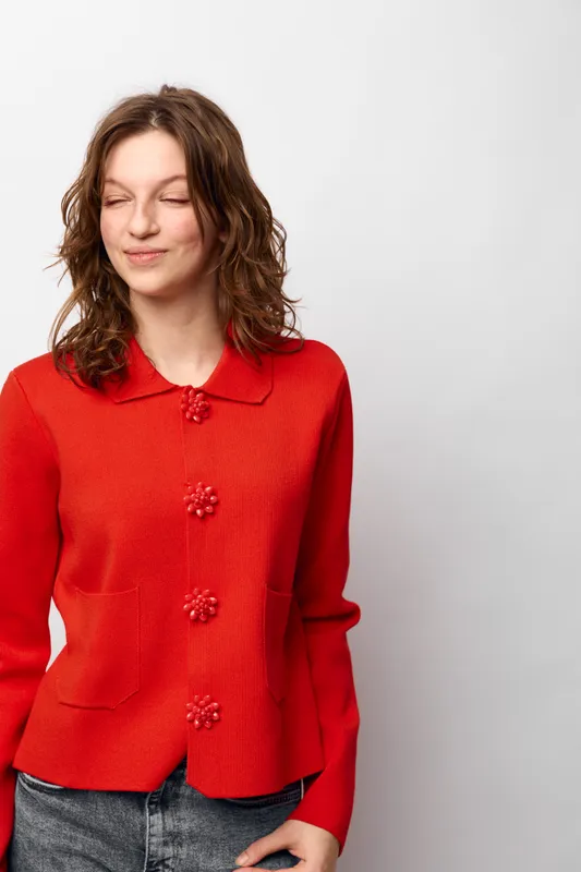 Young woman with curly brown hair wearing a red sweater and jeans stands against white background with eyes closed and slight smile. Casual style and relaxed mood concept.