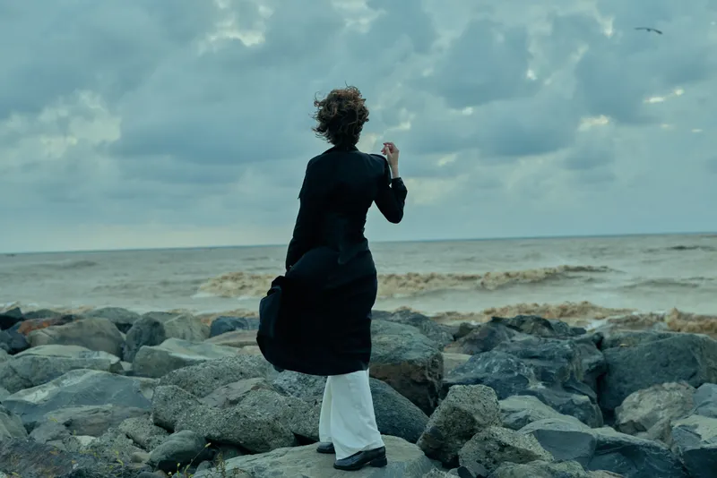 Sea, rocks and wind surround a woman standing on a rugged shore, wearing a dark coat, facing the horizon, conveying resilience, solitude and natural drama on a moody, cloudy day