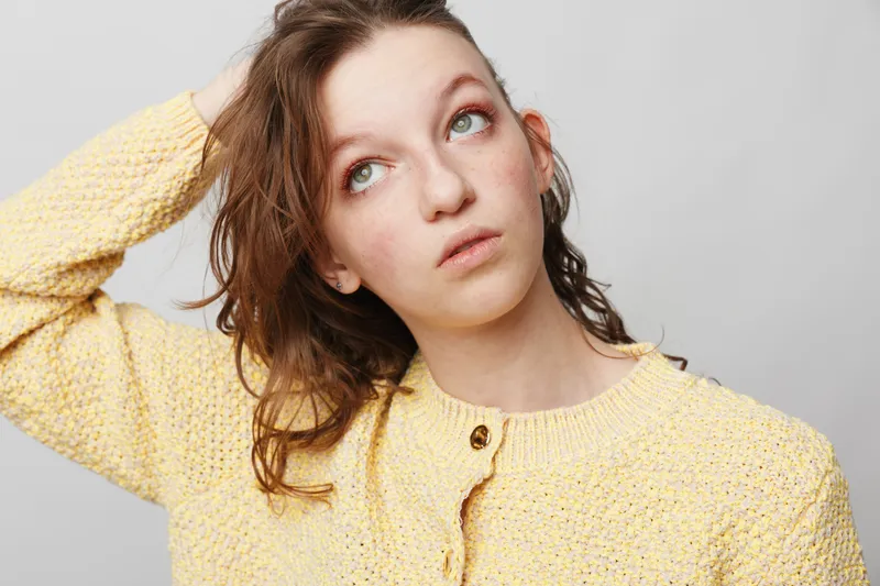 Young woman in yellow sweater looks up thoughtfully with hand in hair against white background. Concept of contemplation, casual style, and natural beauty portrait.
