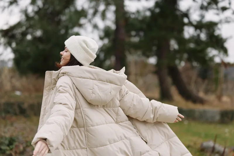 Woman in a beige puffer coat and beanie enjoys a breezy outdoor moment, arms open, in a park with trees and soft autumn light.