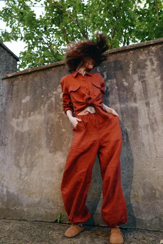 Vibrant woman outdoors in a burnt orange jumpsuit, standing against a weathered wall, wind in hair, relaxed pose, natural light, urban backdrop, candid lifestyle moment