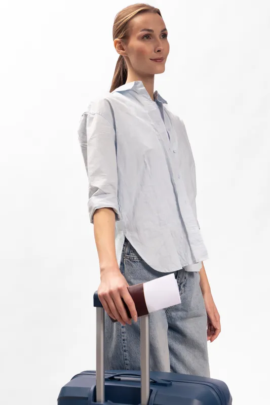 Young woman traveler holding passport and boarding pass stands with suitcase in casual clothes against white background. Concept of travel, vacation, and summer holiday preparation.