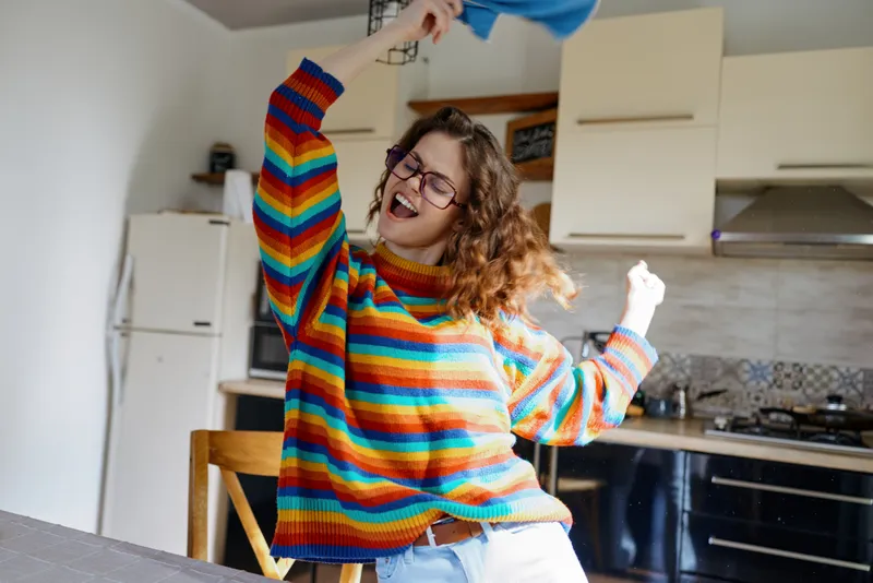 A young woman dances joyfully at a kitchen table, wearing a bright striped sweater, capturing a candid moment of playful home life, with loose hair, glasses on, and sunlight filling the modern space.