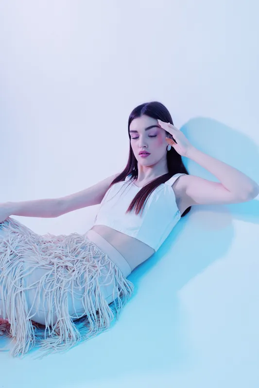 Young woman in white crop top and fringed skirt poses with eyes closed in soft blue light. Fashion concept highlighting beauty, style, and modern elegance in studio setting.