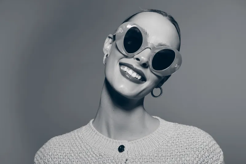 Smiling black woman with dark skin wearing large round sunglasses and hoop earrings in studio portrait.