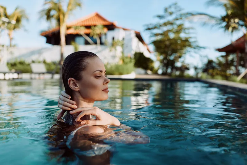 Relaxed woman in a sunlit pool, serene expression, shoulders above water, gentle breeze, tropical backdrop, calm mood, mindful moment, natural colors, vacation vibe, outdoor scene, poolside leisure