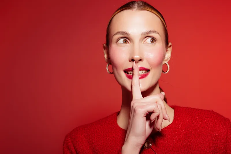 Young woman in red sweater with finger on lips makes silence gesture. She looks sideways with bright lipstick and hoop earrings on red background. Concept of secrecy and quiet mood.