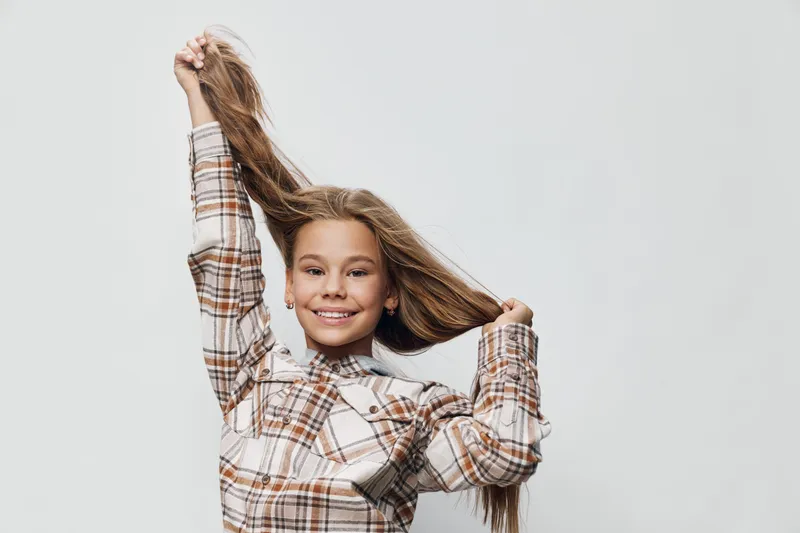 Girl smiling and holding long hair, wearing plaid shirt and white turtleneck against light background, playful mood and casual style portrait