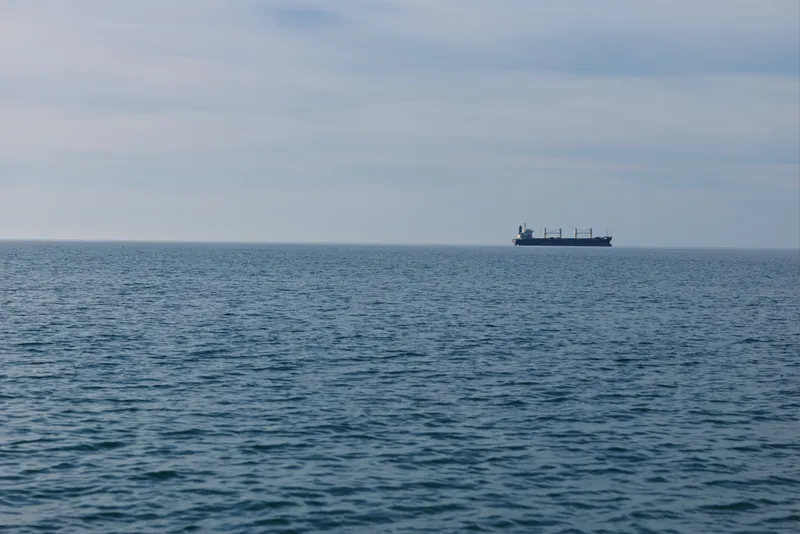 Sea and calm ocean meet at the horizon as a distant cargo ship glides across blue water, a maritime scene highlighting industry, transport and vast open sea.