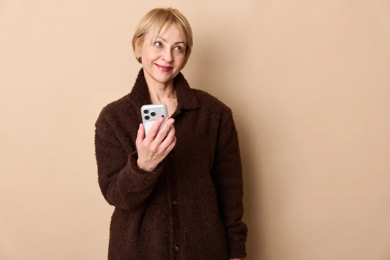 Elderly woman holding smartphone, smiling at camera, beige background, studio portrait. Cheerful communication and everyday technology use, calm indoor lifestyle.