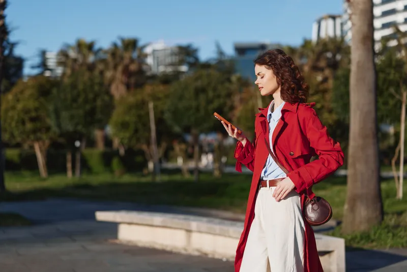 A woman in a red coat walks outdoors, looking at her smartphone, in a sunny park with trees and distant buildings, showcasing casual urban style and confident, relaxed posture