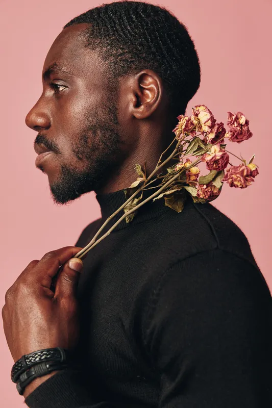 Black man with dark skin holding dried flowers on shoulder in creative conceptual visual style. Portrait with thoughtful expression and artistic mood on pink background in studio.
